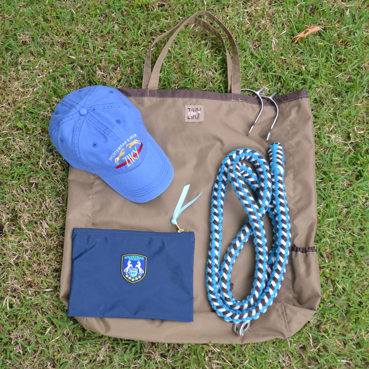 Taupe ZIKY tote bag lifestyle photo with blue equestrian cap, blue crest cosmetic case, horse lead rope, and stirrups on green grass